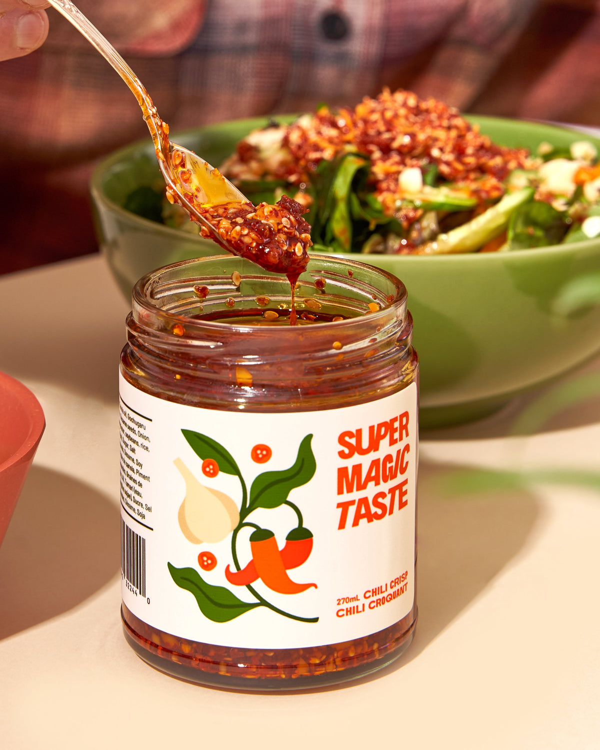 A spoonful of Super Magic Taste chili crisp lifted from the jar, showing the crunchy chili and sesame solids, with a salad bowl behind.