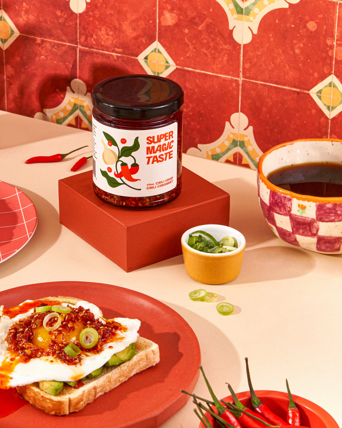 A slice of avocado toast topped with a fried egg and Super Magic Taste chili crisp, the jar behind it on a red tiled counter.