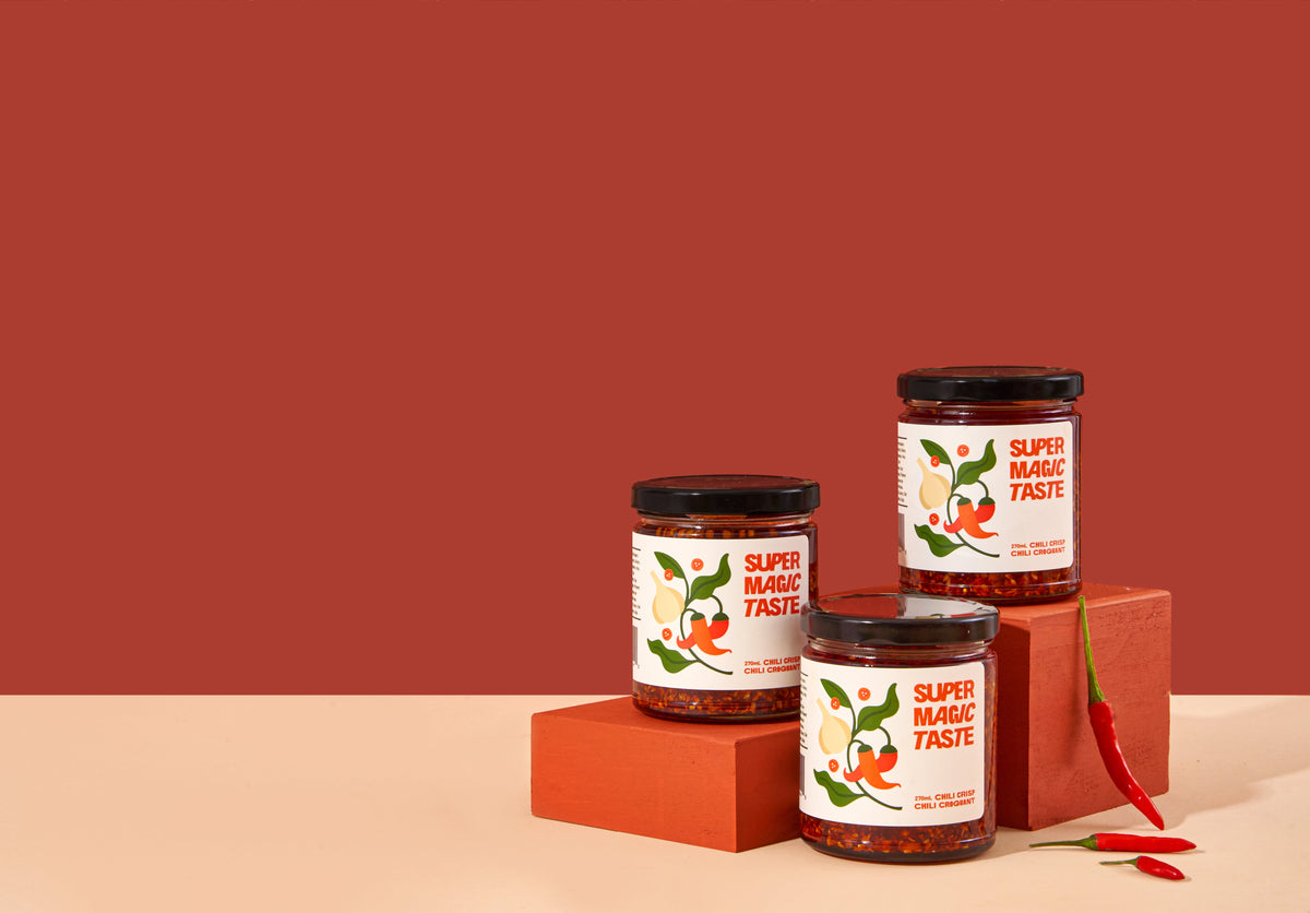 Three jars of Super Magic Taste chili crisp arranged on terracotta blocks against a deep red background.
