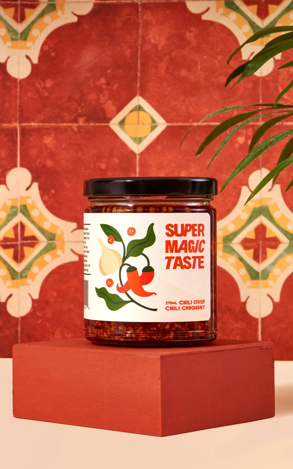 A jar of Super Magic Taste Chili Crisp on a terracotta plinth, the crunchy chili, garlic and almond bits visible through the glass.