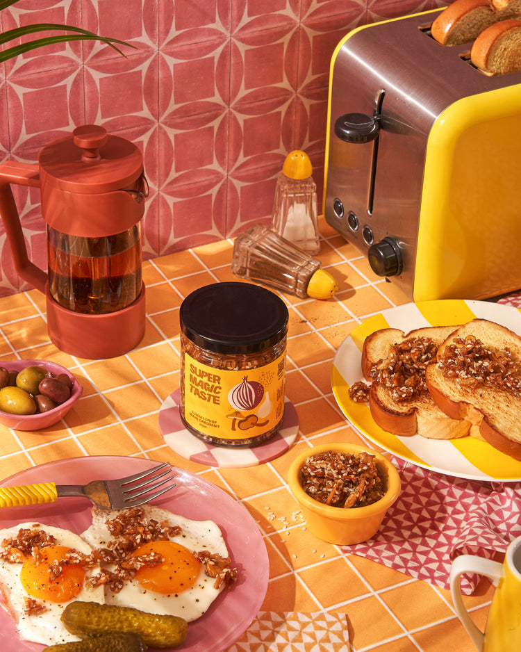 Super Magic Taste Yummy Crisp spooned over fried eggs and buttered toast on a yellow breakfast table, with the jar in the middle.