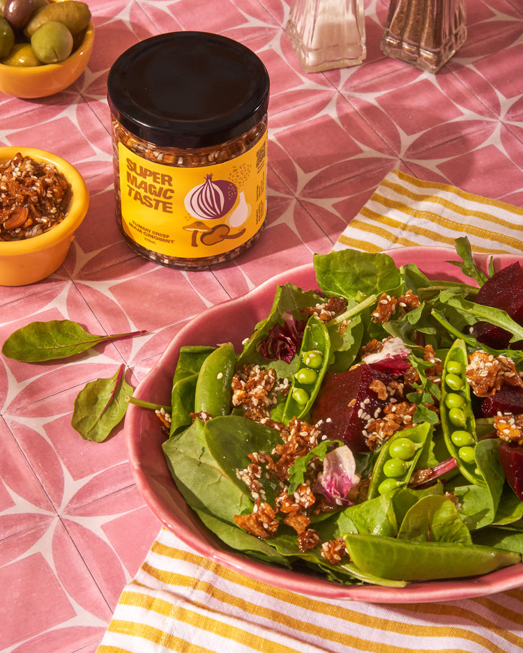 A spinach and beet salad scattered with Super Magic Taste Yummy Crisp, the jar behind it on a pink cloth.