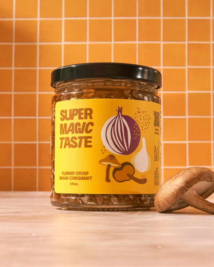 A jar of Super Magic Taste Yummy Crisp mushroom chili crisp on a counter against orange tile, with a shiitake mushroom beside it.