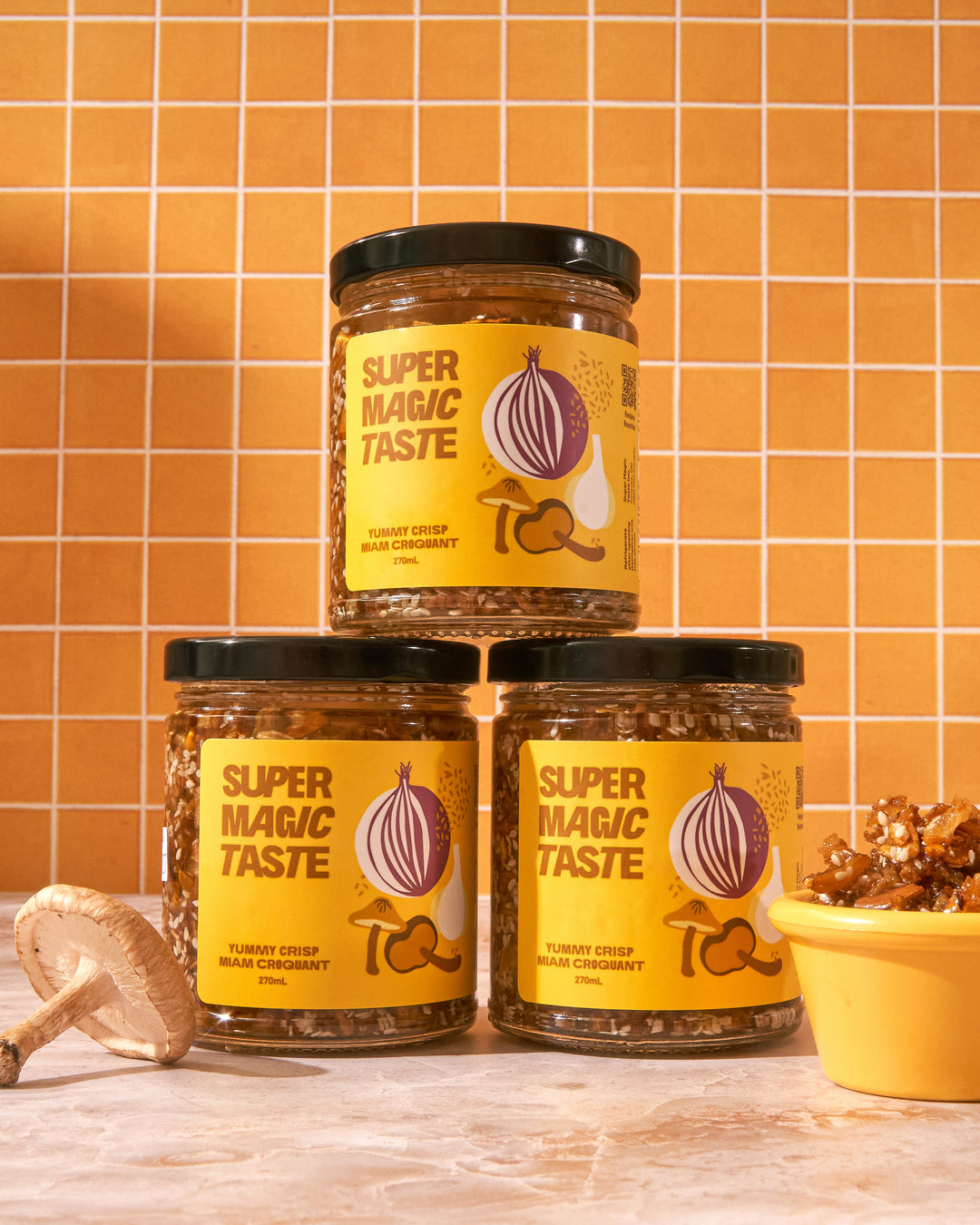 Three jars of Super Magic Taste Yummy Crisp stacked against orange tile with shiitake mushrooms and a dish of the crisp.