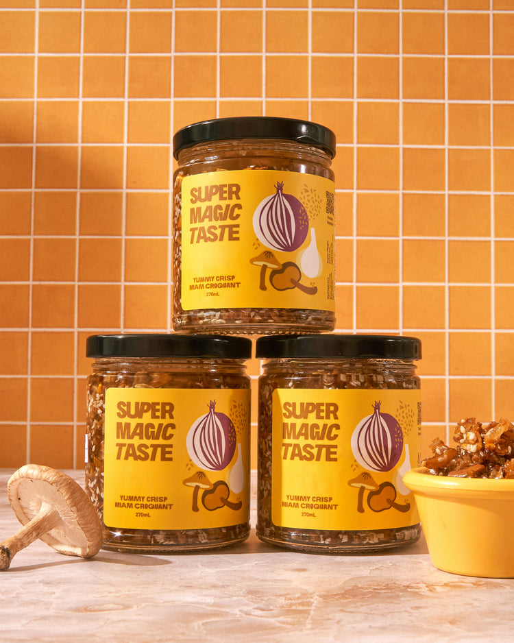 Three jars of Super Magic Taste Yummy Crisp stacked against orange tile with shiitake mushrooms and a dish of the crisp.