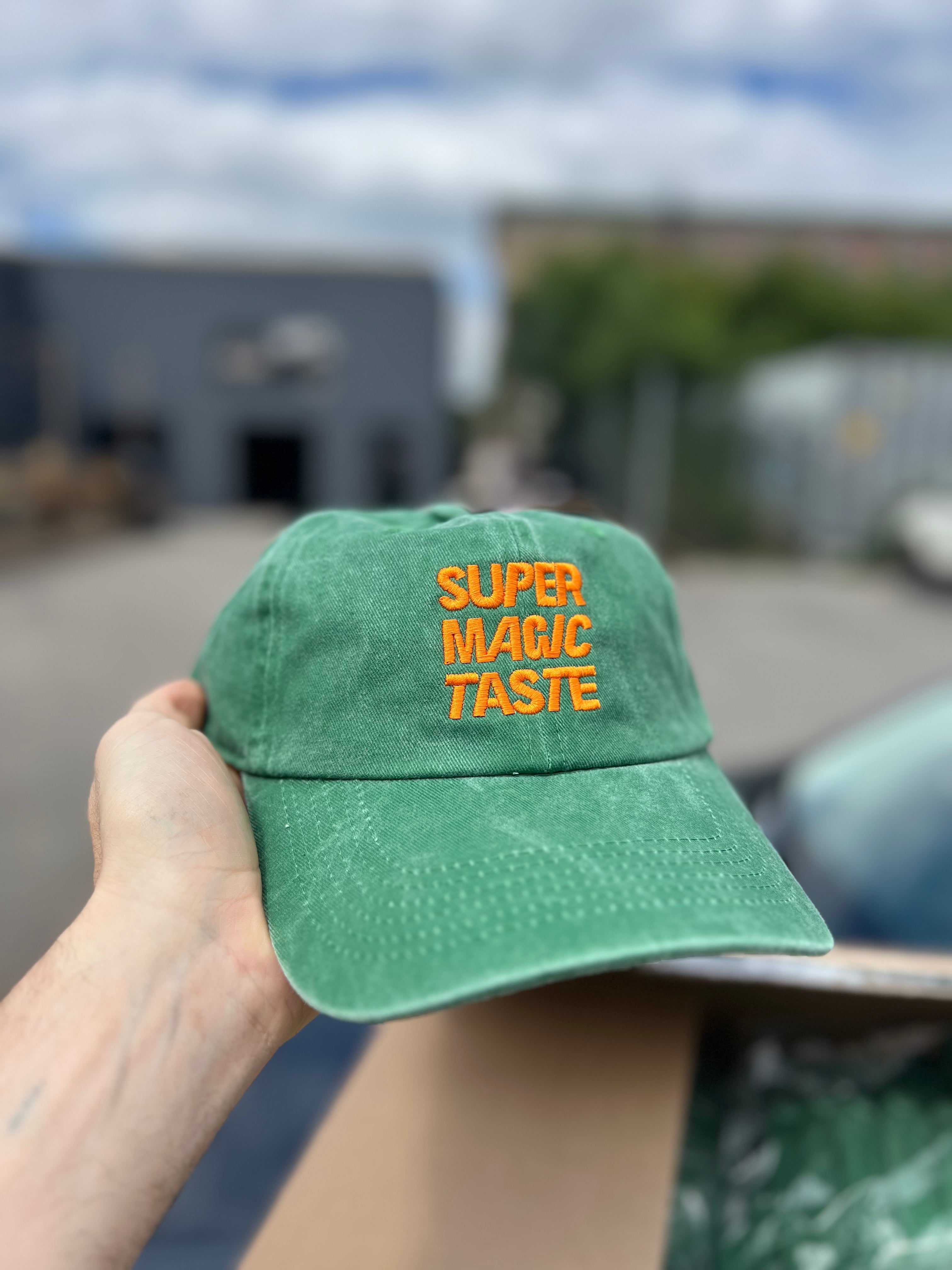 A hand holding up a green Super Magic Taste cap with the logo embroidered in orange