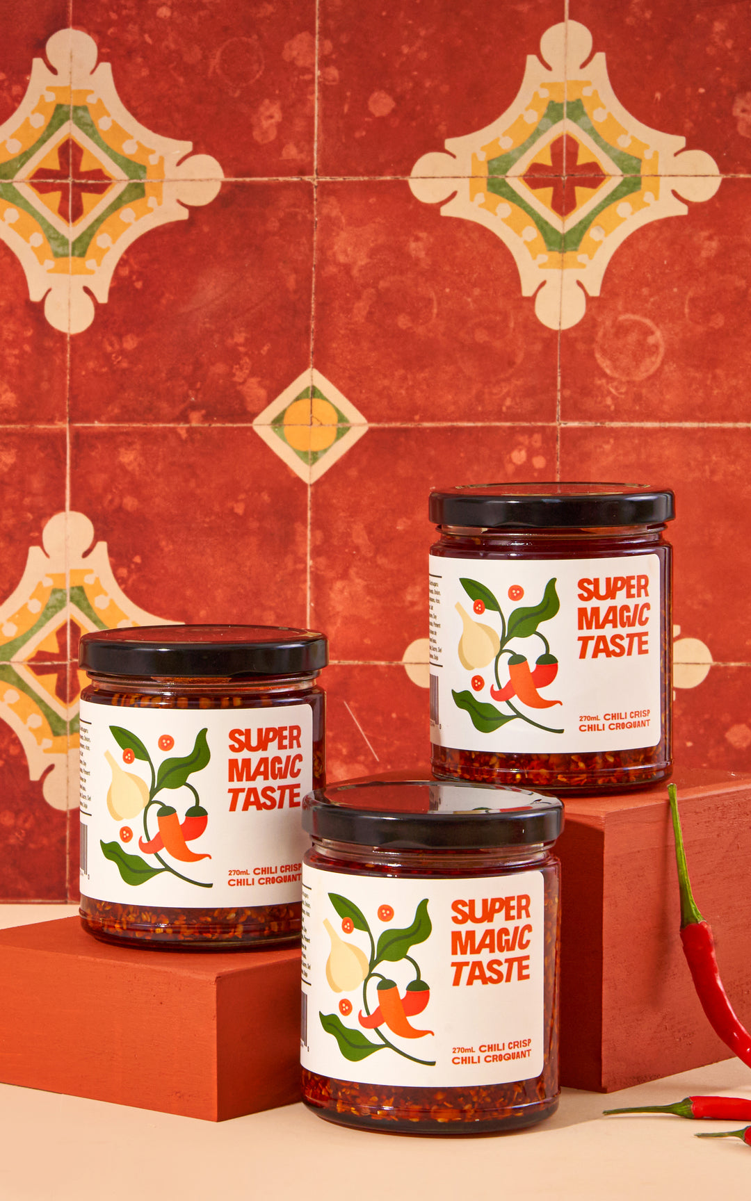 Three jars of Super Magic Taste Chili Crisp stacked on terracotta blocks against red patterned tile, with fresh red chilies.